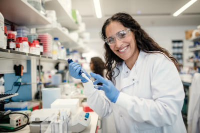 A scientist in a lab coat is in a laboratory. working. She is smiling at the camera.  Image from Alzhiemer's Research UK.