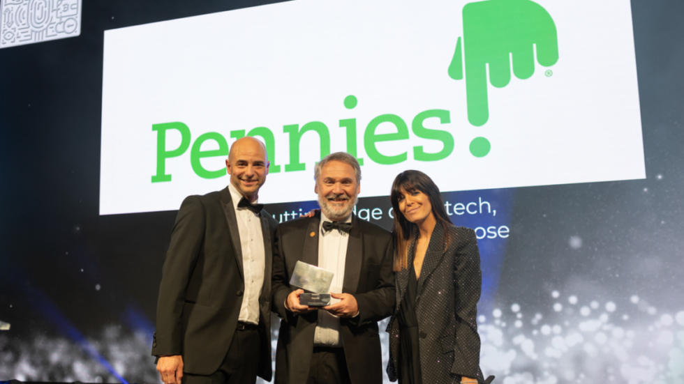Paul Seaman accepts the Industry Achievement Award at the Card & Payments Awards 2022 on behalf of Pennies, from host Claudia Winkleman and Rupert Bedell, Vice President of Marketing at American Express