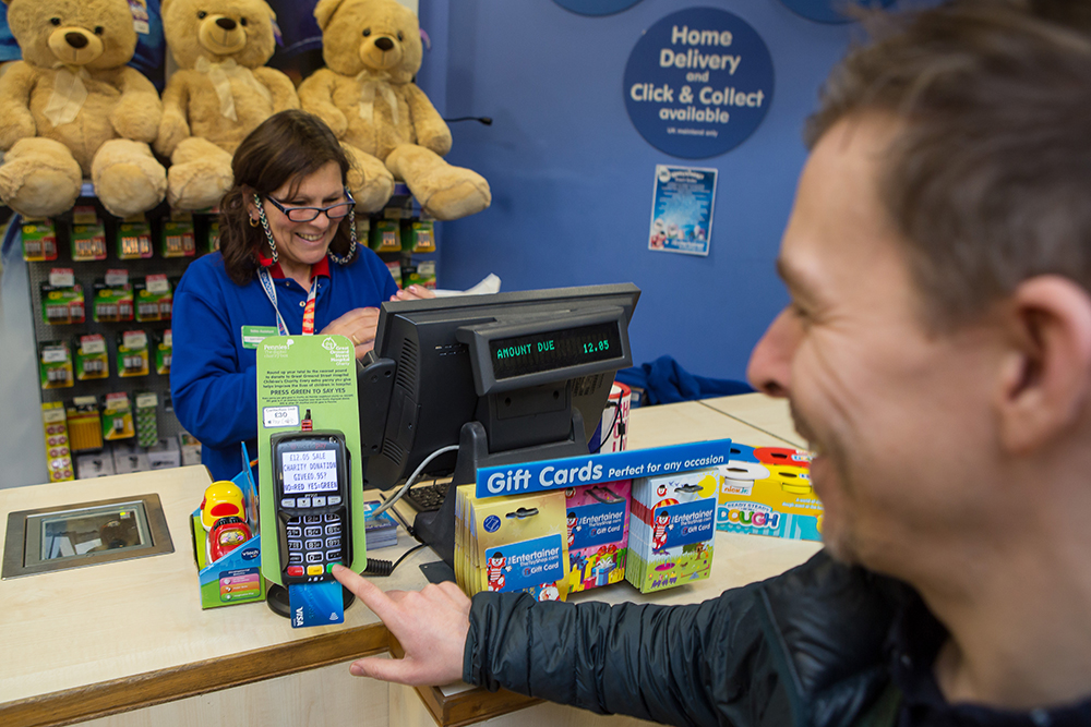 A customer uses a card payment machine to donate in-store at The Entertainer toy shop. The point of sale material is attached to the card machine, clearly displaying Great Ormond Street Children's Hospital Charity, alongside the Pennies logo. The shop assistant and customer share a smile.